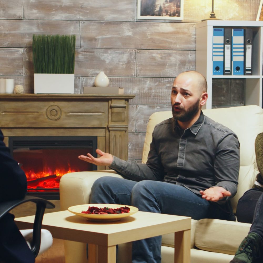 Woman sitting on couch crying at couples therapy while the husband is yelling at therapy.